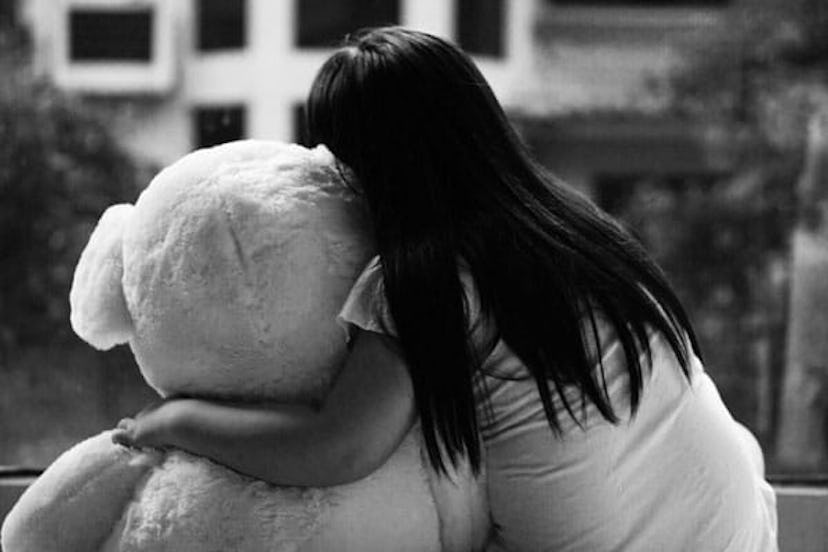 The back of a dark-haired girl with long hair in a white shirt hugging a huge teddy bear in black an...