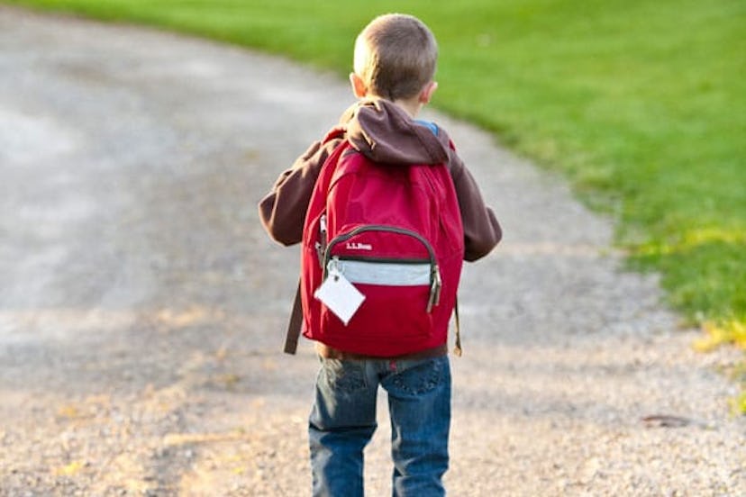 The back of a boy in a brown hoodie and blue denim jeans carrying a red backpack on his back while w...