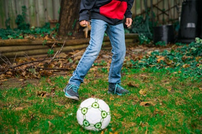 A boy in a black-and-red hoodie and denim jeans playing football alone in a backyard