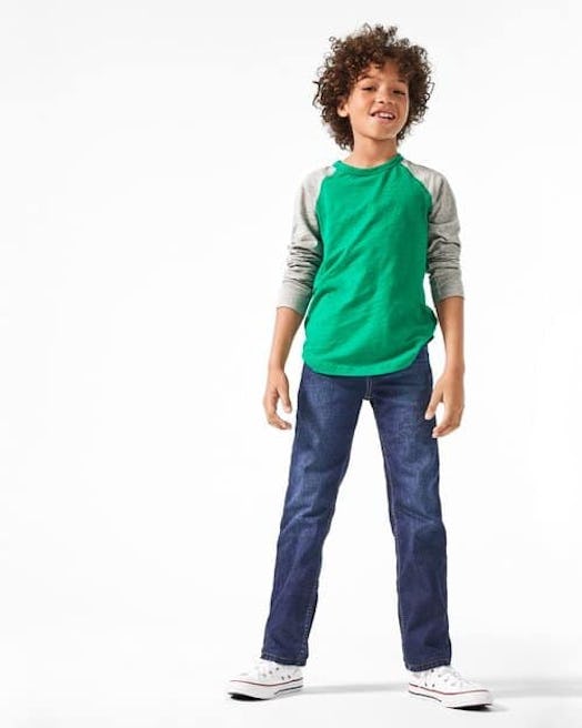 A curly-haired boy in a green shirt with gray sleeves, blue denim jeans, and white all-star sneakers...