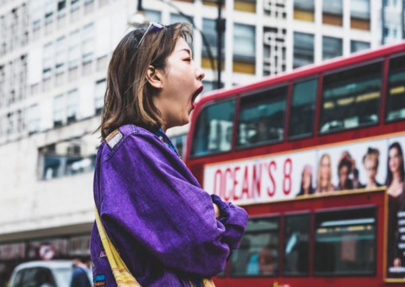 A woman in her 30s standing on a street and yawning 