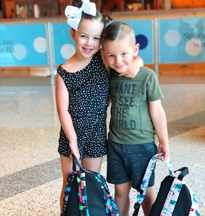A sister hugging her brother while they’re smiling, holding backpacks in their hands