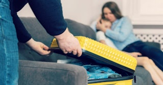 Man closing a yellow suitcase on a grey couch with a woman holding a child in the back