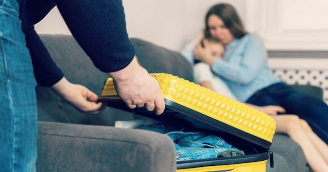 Man closing a yellow suitcase on a grey couch with a woman holding a child in the back