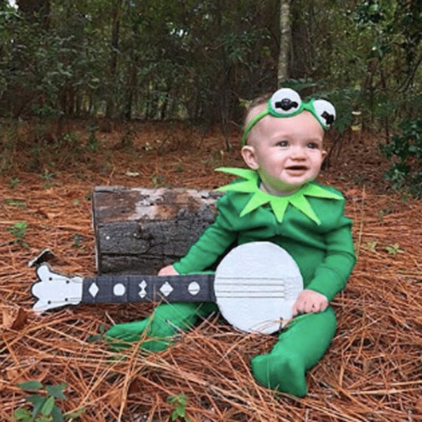 A baby sitting in a forest in a Kermit The Frog Halloween costume 
