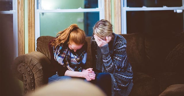 A red-haired and a blonde woman sitting on a couch with their heads down, holding hands