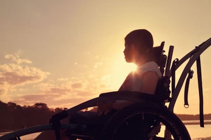 A preteen girl sitting in a wheelchair next to a river, watching the sunset.