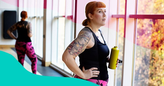 A woman with internalized fat phobia in a black top and pink-black pants standing in a gym and holdi...