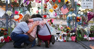 Two brunette women and a blonde woman kneeling in front of a Parkland shoot victims memorial wall.