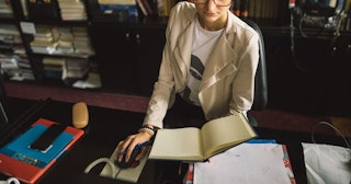 A school secretary wearing a white shirt, glasses and a coat sitting at her table holding a book and...
