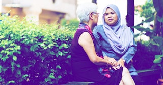 An elderly grey hair woman in a sleeveless dress comforting a woman with a lilac hijab, and they're ...