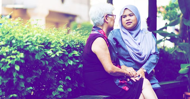 An elderly grey hair woman in a sleeveless dress comforting a woman with a lilac hijab, and they're ...