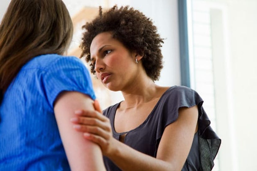 A short-haired curly woman in a short-sleeved grey shirt comforting and holding the arms of her frie...