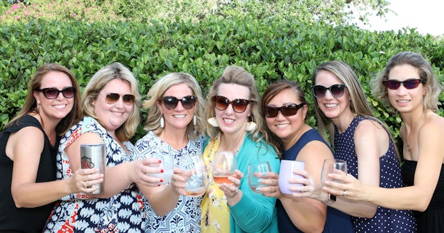 Seven friends posing outside with sunglasses on and with drinks in their hands while smiling