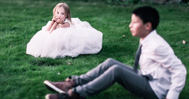 A girl in a white dress and a boy in a suit sitting on the grass who are indoctrinated to the cisgen...