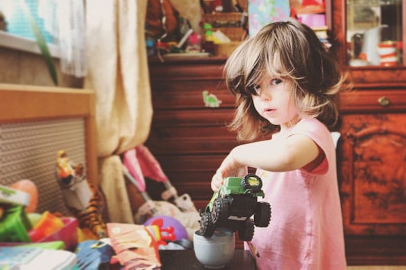 Little girl in pink dress in a room full of toys. She is placing a toy truck in a white coffee cup