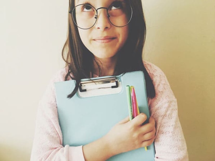 Young girl wearing round glasses and a light pink sweater. She is looking up and holding a clipboard...