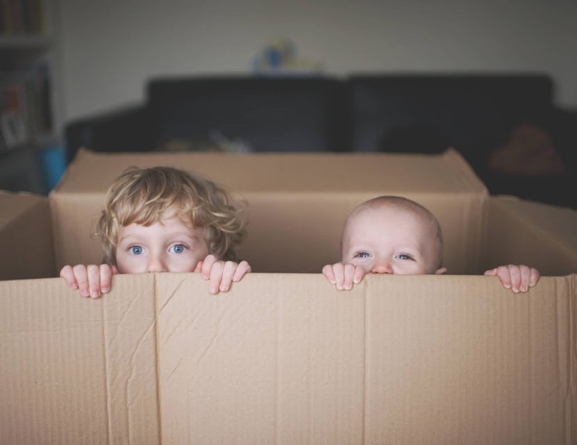 Small kid and baby playing in and looking out of a cardboard box.