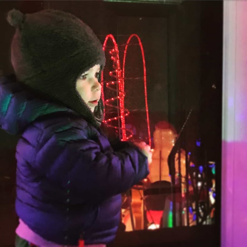 A toddler wearing a winter jacket and a hat standing next to a window