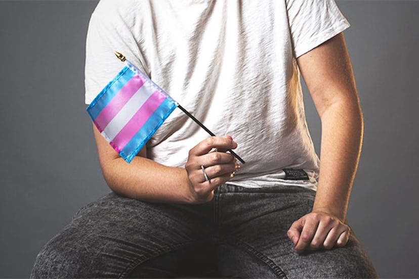 A young person in a white t-shirt and black pants sitting while holding a transgender flag in their ...