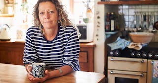 A sober, blonde, mature woman leaning on the kitchen table while holding a cup, looking toward the c...