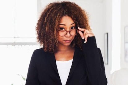 A curly-haired brunette woman in a white shirt and black blazer lowered glasses with a judgmental lo...
