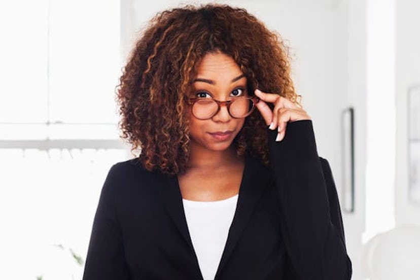 A curly-haired brunette woman in a white shirt and black blazer lowered glasses with a judgmental lo...