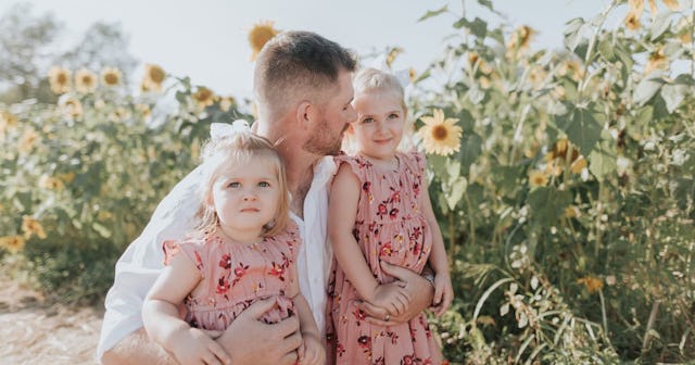 A dad in a white shirt with his two daughters in matching pink floral dresses in a sunflower field