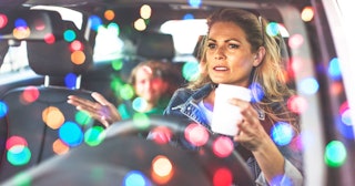 A mom in a car with her kid surrounded by Christmas lights that are reflecting on her car’s windshie...