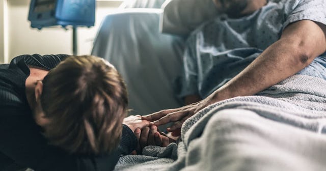 A man in a hospital bed in a blue T-shirt covered with a white blanket and a woman at the edge of th...