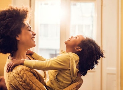 A curly black-haired woman in a beige sweater holding her daughter up and laughing with a window in ...