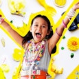 A small girl smiling with her hands up surrounded by yellow plastic trinkets and junk