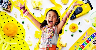 A small girl smiling with her hands up surrounded by yellow plastic trinkets and junk