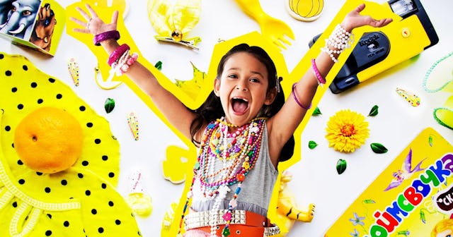 A small girl smiling with her hands up surrounded by yellow plastic trinkets and junk