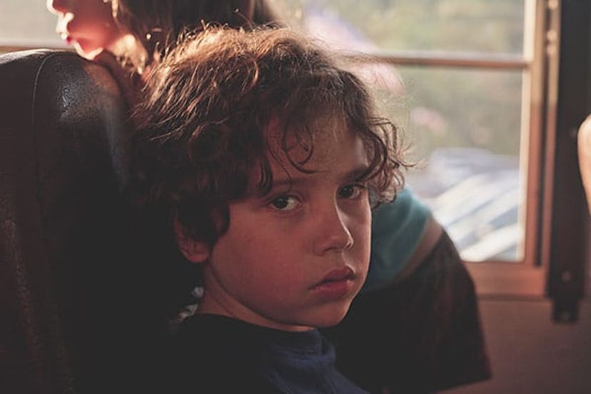 A child sitting on the bus and looking at the camera; a child next to him standing up on the bus sea...