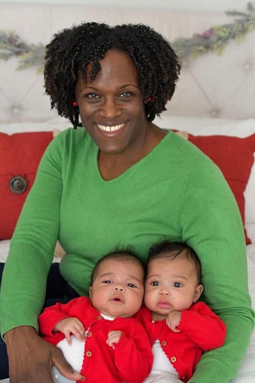 A smiling mother in a green sweater holding her twin babies in red shirts