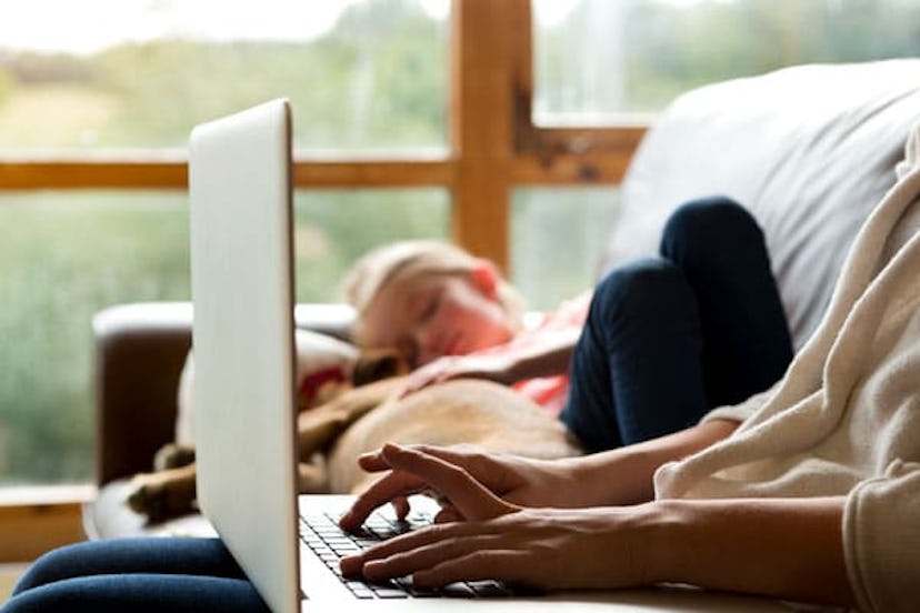 A working mom in a cream sweater typing on her laptop in her lap on a couch next to a sleeping child...