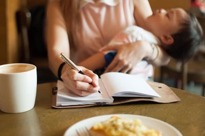 A working mom in a pink top with a white collar holding her baby and writing something down in a not...