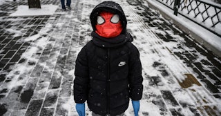 boy wears a Spiderman mask over his protective mask and rubber gloves while standing in the street