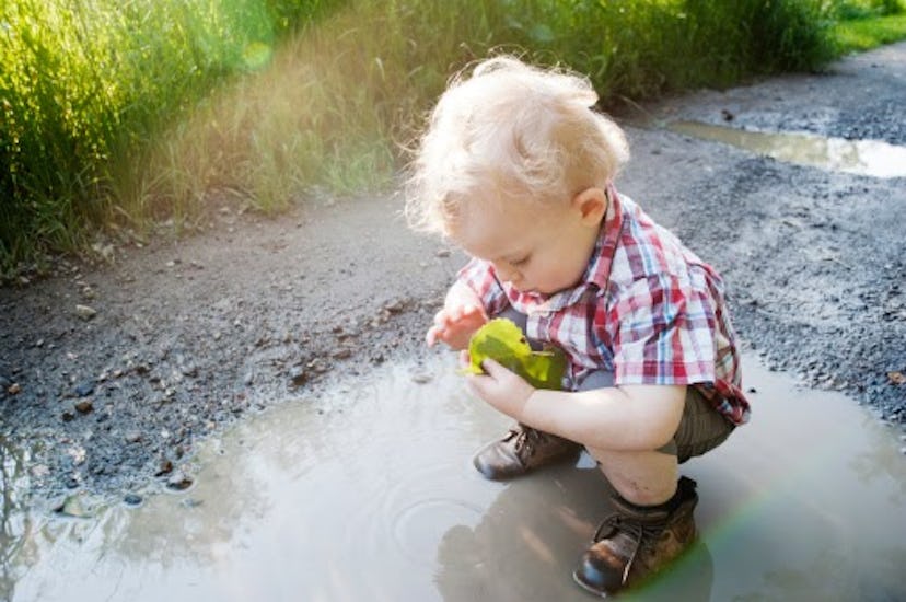A toddler boy is standing in a muddy puddle inspecting a leaf he is holding in his hand