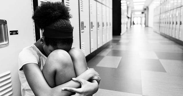 Sad preteen girl hides her head in her knees while sitting in a school hallway or locker room.
