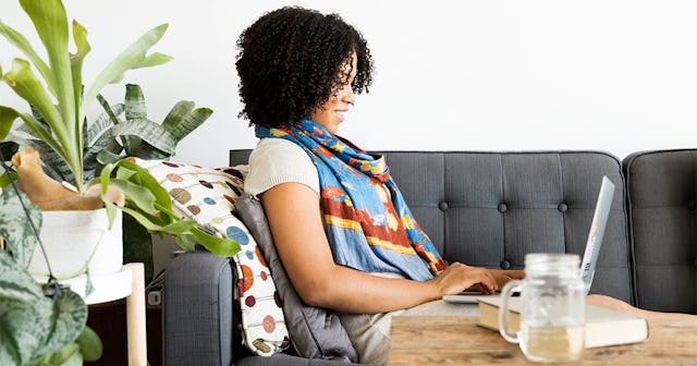 A woman smiles as she relaxes on a stylish gray sofa, working on her laptop. Potted plants flourish ...