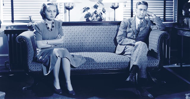woman sits on one side of a couch folding her arms and wearing an angry expression while a man sits ...
