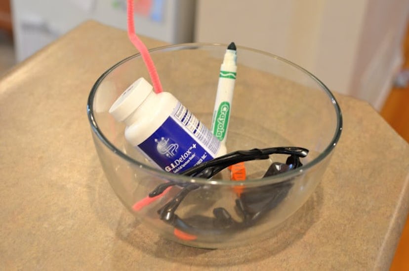 A glass bowl on a brown wood surface filled with a toothbrush, sunglasses, medicine and a green mark...