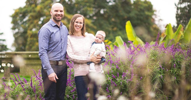 Husband, wife, and baby smiling and posing outside surrounded by flowers