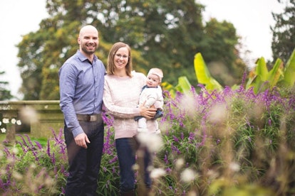 Husband, wife, and baby smiling and posing outside surrounded by flowers