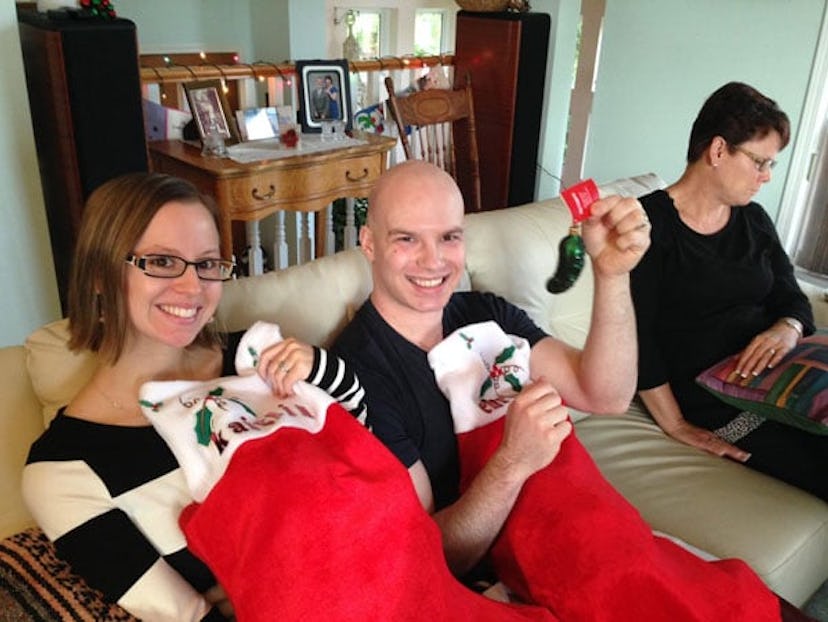 Bald husband with cancer sitting on a beige sofa, holding Christmas stocking bags and smiling with h...