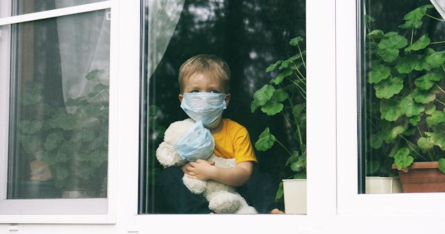 Sad child and his teddy bear both in protective medical masks sits on windowsill and looks out windo...