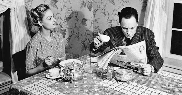A woman, looking angry at being ignored, sits with her husband as reads the newspaper over breakfast...