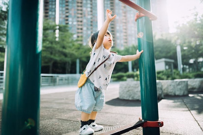 Playful little toddler girl arms raised and trying to reach a pull up bar at the outdoor playground